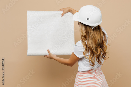 Back rear view young employee laborer handyman woman wearing white t-shirt helmet hold wallpapers isolated on plain beige background Instruments accessories for renovation room. Repair home concept. photo