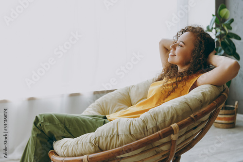 Side view fun young woman of African American ethnicity 20s wear casual clothes sits near window in armchair close eyes dream stay at home flat rest relax spend free spare time in living room indoor