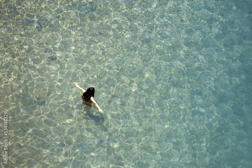 Aerial view to azure sea and woman in bikini go to swim in transparent water. Girl swimmer with arms outstretched, beach vacation