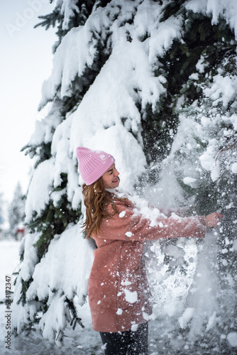 girl is having fun near a snow-covered Christmas tree in a pink jacket and hat, it's snowing