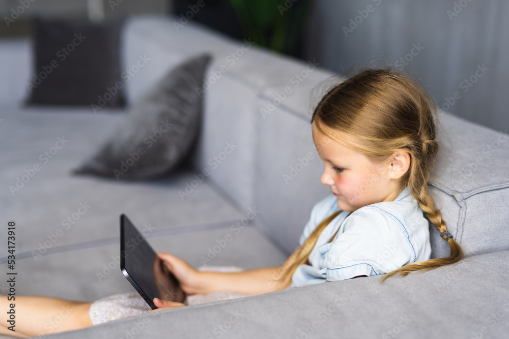 Smiling little girl lying on couch, enjoying playing online game on digital tablet computer. Addicted to technology, happy small kid using funny applications, web surfing information. Smiling little girl lying on couch, enjoying playing online game on digital tablet computer. Addicted to technology, happy small kid using funny applications, web surfing information.