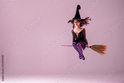 Obraz na plátne A ballerina on pointe shoes in a black witch costume in a hat flies on a broomstick on a lilac background