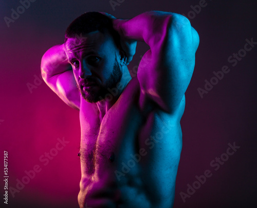 Sportive man bodybuilder is posing in the colorful neon light with naked muscular torso showing chest, abdominal muscles in neon studio light.