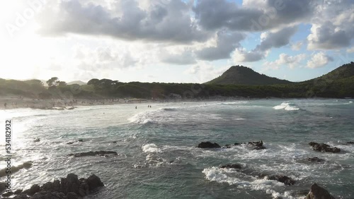 The waves splashing on beach coastline of Cala Agulla (Cala Ratjada) on Mallorca in the mediterranean sea. Deep blue water and a green mountain in the background. People bathing, surfers surfing. 4k.