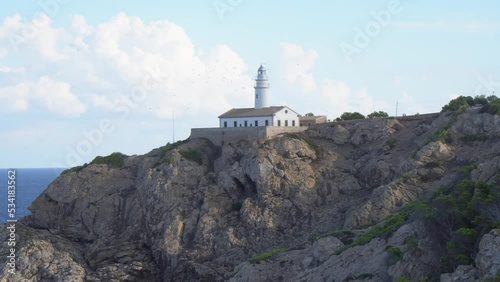 The lighthouse of Cala Ratjada. Waves splashing on a rocky coastline of Mallorca in the mediterranean sea. Deep blue water and steep cliffs. A flock of birds flying. 4k.