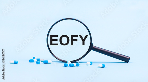 On a blue background, white and blue capsules with pills and a black magnifying glass with the text EOFY. Medical concept.