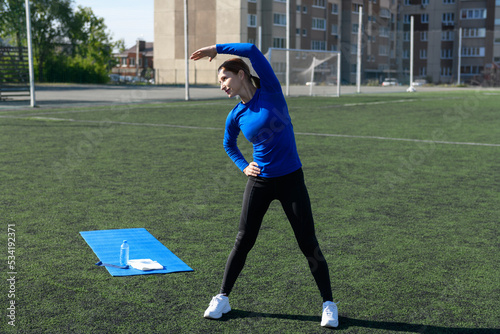 Women and sport. Girl in sportswear does exercises: bends and stretches on the grass at the stadium on a sunny day. Middle aged sportswoman dressed in sportsclothes exercising outdoors photo