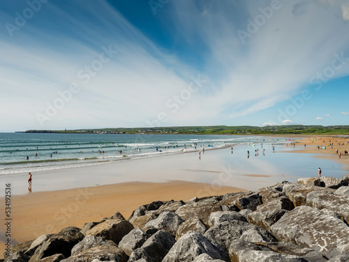 Yellow sand and blue ocean and sky. People swim in the background and surf on board. Lahinch beach in county Clare, Ireland. Warm sunny day. Irish landscape.