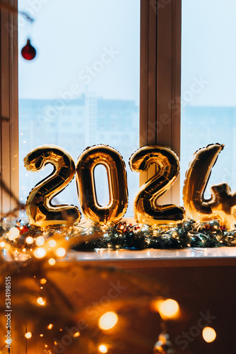 2024 golden foil balloons on blue window sill. Celebrating holidays at home, festive decor concept. Happy New Year 2024. close-up numbers of year 2024 on dark background. Bokeh warm garland light.