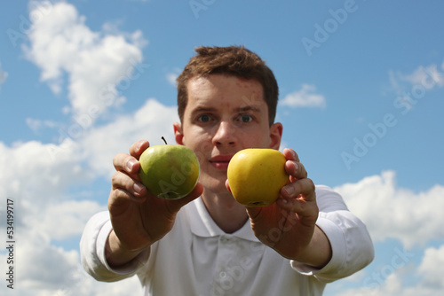 Young man with green and yellow apple on a blue sky background. Pavel Kubarkov, two apples in my hands and sky. Photo was taken 16 July 2022 year, MSK time in Russia.