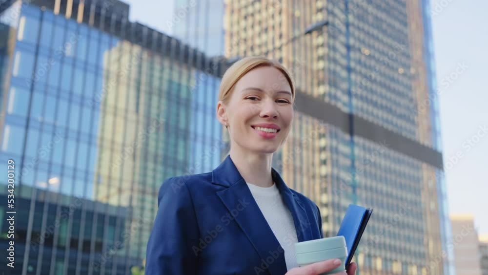 Happy business woman looking at camera and smiling standing outdoors near modern office building in city. Successful manager in blue formal suit holding documents folder and coffee cup in downtown Happy business woman looking at camera and smiling standing outdoors near modern office building in city. Successful manager in blue formal suit holding documents folder and coffee cup in downtown