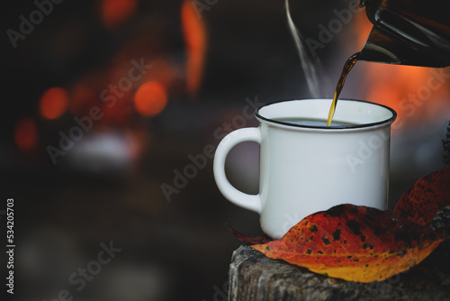Cuadro en lienzo Kettle pouring hot steaming coffee into a white enamel cup  sitting on an old log with autumn leaves