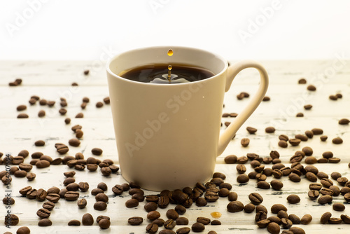 A mug of hot coffee with evaporation and steam and a small splash on a wooden table next to coffee beans