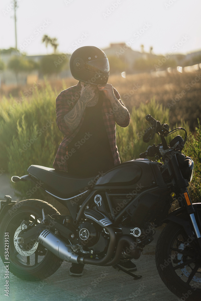 Fototapeta premium Chico joven tatuado con camisa a cuadros roja montando motocicleta por carretera solitaria al atardecer