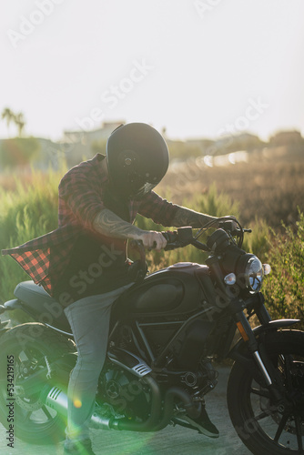 Chico joven tatuado con camisa a cuadros roja montando motocicleta por carretera solitaria al atardecer