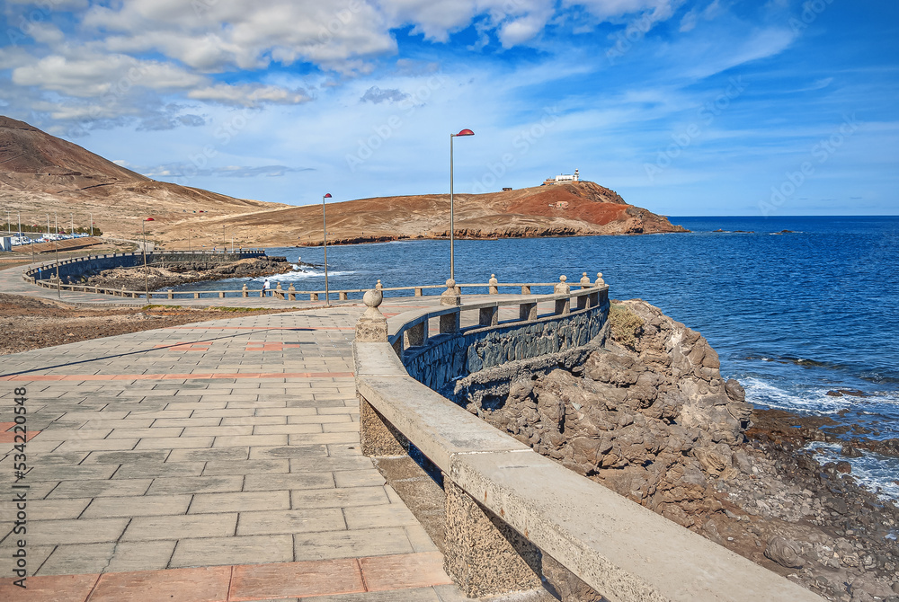 Empty stone embankment. A view of the ocean and a small lighthouse at the entrance pier of the island. Calm blue sea and sky. Empty stone embankment. A view of the ocean and a small lighthouse at the entrance pier of the island. Calm blue sea and sky.