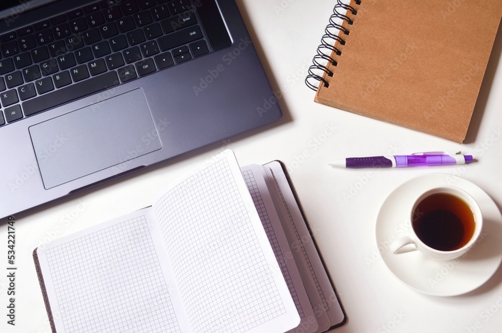 Office table top view stock photo. Laptop, open paper notebook, purple pen and cup of coffee. Workplace flat lay image, business background Office table top view stock photo. Laptop, open paper notebook, purple pen and cup of coffee. Workplace flat lay image, business background