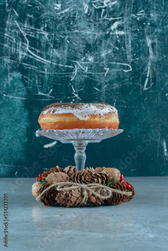 Glazed donut on a glass pedestal in the middle of a pine wreath on marble background