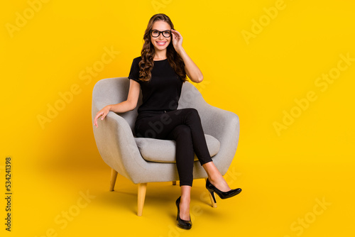 Photo of adorable thoughtful young lady dressed black clothes eyewear sitting grey chair smiling isolated yellow color background photo