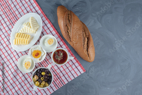 Tablecloth underneath a breakfast bundle of bread loaf and platters of cheese, butter and egg, with tea and olives on marble background photo