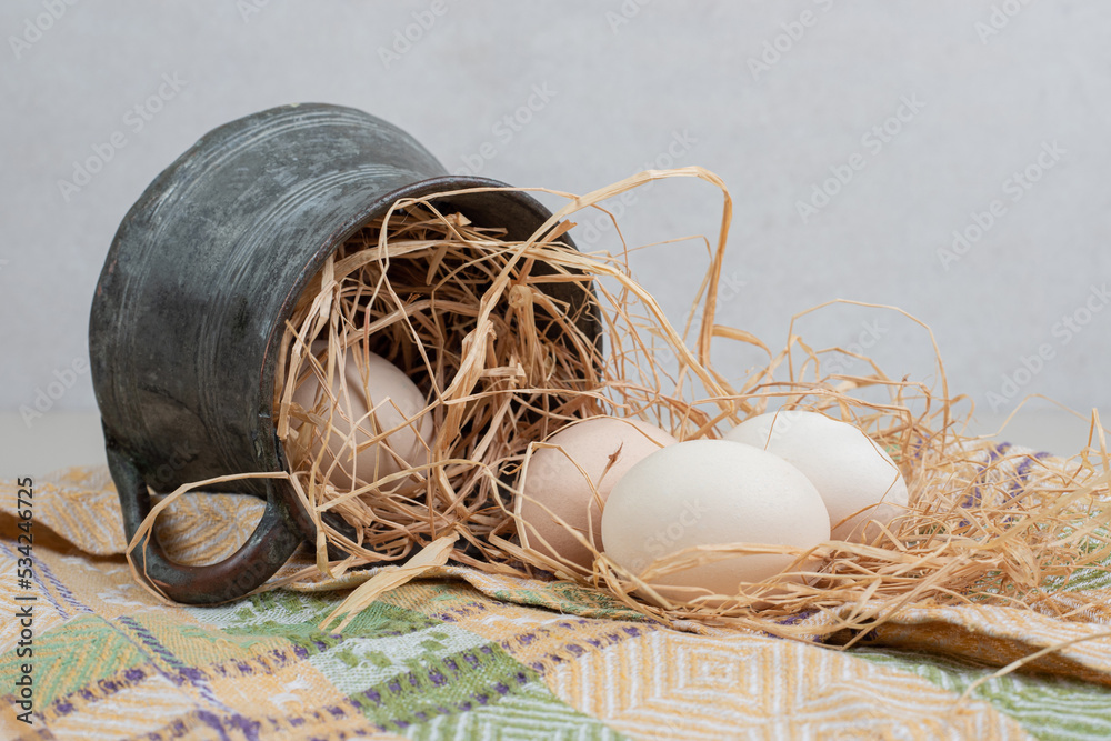 Fresh chicken eggs with hay in ancient cup on tablecloth