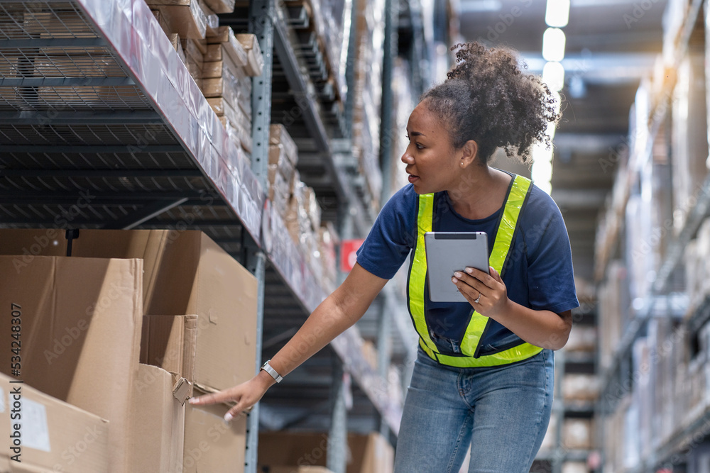 African-American working woman carries a tablet while counting inventories and picking items from shelves to deliver logistics to customers in a warehouse.