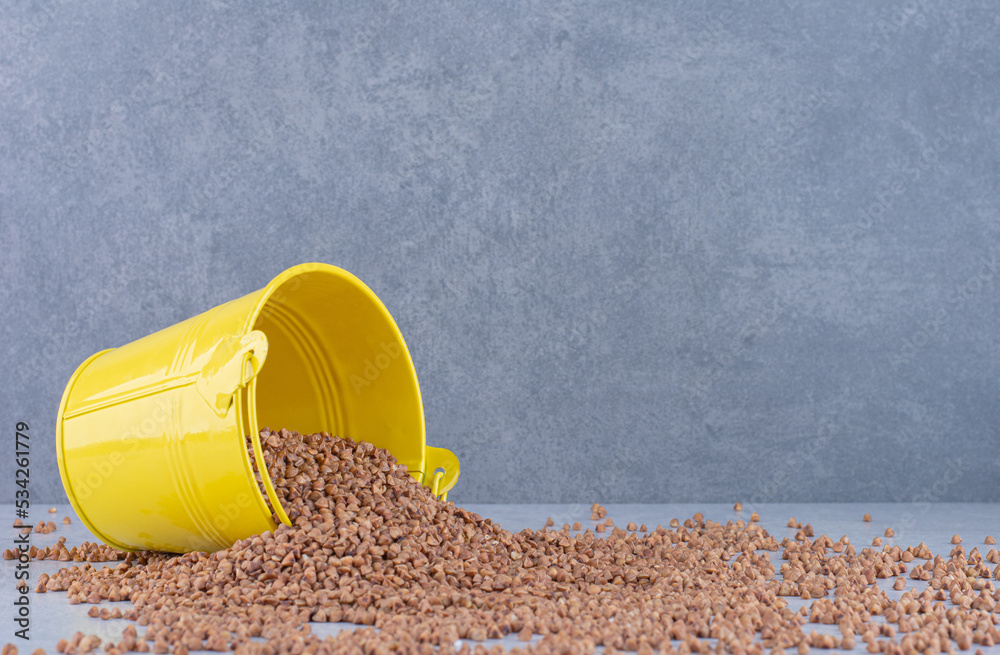 Small bucket fallen over on top of a heap of scattered buckwheat on marble background Small bucket fallen over on top of a heap of scattered buckwheat on marble background
