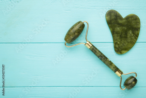 Green jade roller and gua sha stone for facial massage and on blue background. Home beauty and selfcare accessories. Face roller for anti age wrinkle treatment. Top view, flat lay. photo