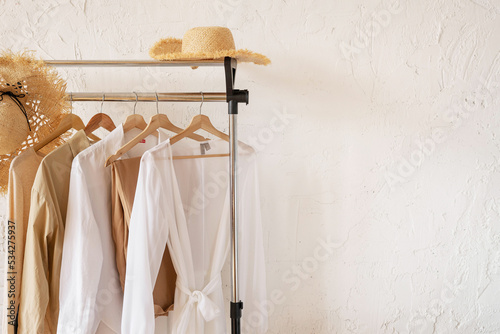 Trendy capsule wardrobe in beige and white on a rail rack with straw hats. Copy space
