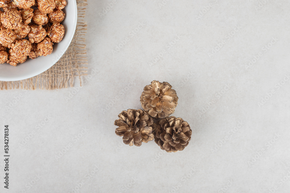 Three conifer cones next to a small bowl of popcorn candy on marble background Three conifer cones next to a small bowl of popcorn candy on marble background