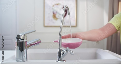Child’s hands in the kitchen draw filtered water into a pink cup. Water flows from a small filter faucet. The filter faucet is in the center, medium shot.4K