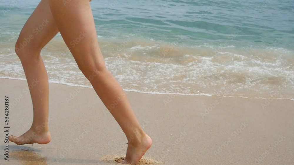 Bare feet of young woman jogs over wet sand in shallow waterline. Female legs runs along seashore. Girl spends time near ocean coast. Summer vacation or holiday concept. Close up Slow motion Bare feet of young woman jogs over wet sand in shallow waterline. Female legs runs along seashore. Girl spends time near ocean coast. Summer vacation or holiday concept. Close up Slow motion