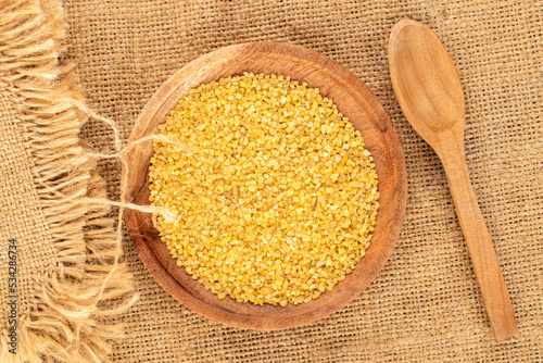 Konstfotografi Raw bulgur grains in a wooden plate with a spoon on a jute cloth, macro, top view