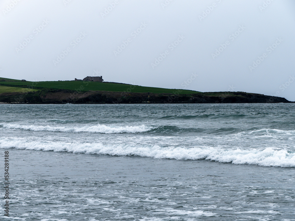 Sea at high tide. A building on a hill. Gray sky, building on green grass field near sea. Seaside landscape, white foam on the waves.