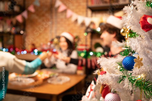 Close-up and focus of a half decorated white Christmas tree on a blurred background of happy friends in Santa hats sitting at a dining table clinking red wine glasses together to celebrate Christmas.