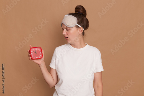 Horizontal shot of sad upset sleepy Caucasian woman wearing white t shirt and sleeping mask looking at red alarm clock, feels sleepy, have lack of sleep, posing isolated over brown background.