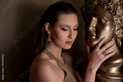 Elegant fashionable brunette woman with make up on clean skin near golden Apollo bust. Beauty revitalize, rejuvenation