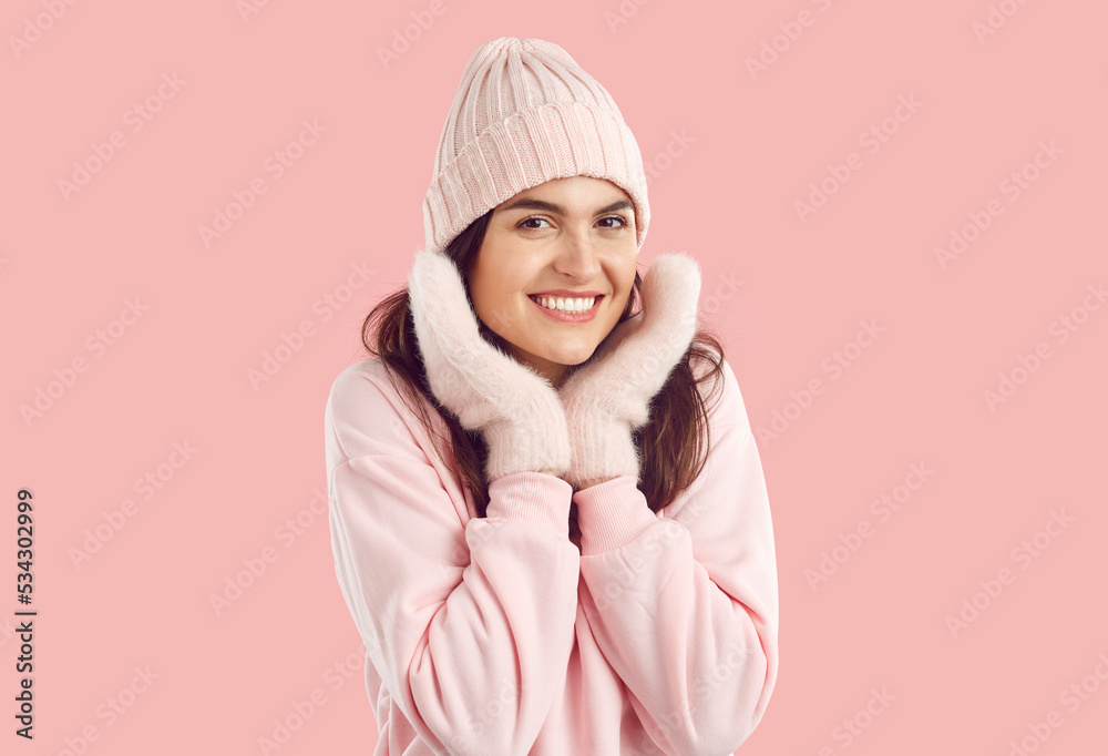 Happy smiling woman in winter mittens and hat holding hands near face on pink background in studio. Gentle cute woman with beautiful smile and wearing light pink winter clothes is looking at camera. Happy smiling woman in winter mittens and hat holding hands near face on pink background in studio. Gentle cute woman with beautiful smile and wearing light pink winter clothes is looking at camera.