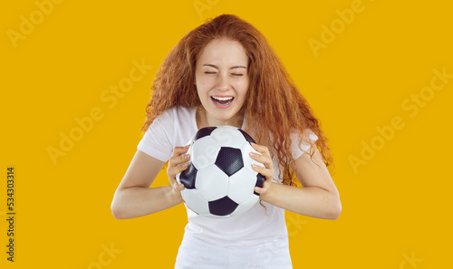 Happy emotional woman holding soccer ball. Beautiful young woman or college girl with long ginger hair standing isolated on yellow background, holding football and laughing with joyful face expression © Studio Romantic