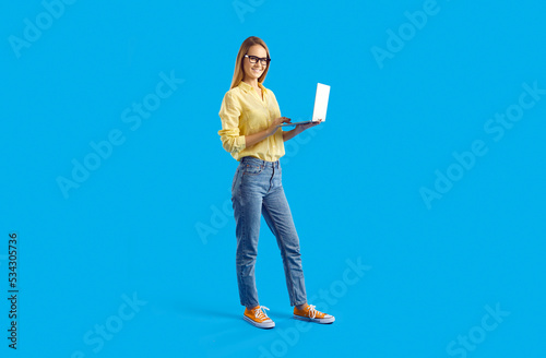 Happy young woman using modern laptop computer. Full body studio shot of beautiful student girl in shirt, jeans and eyeglasses standing against solid blue background, holding notebook PC and smiling photo