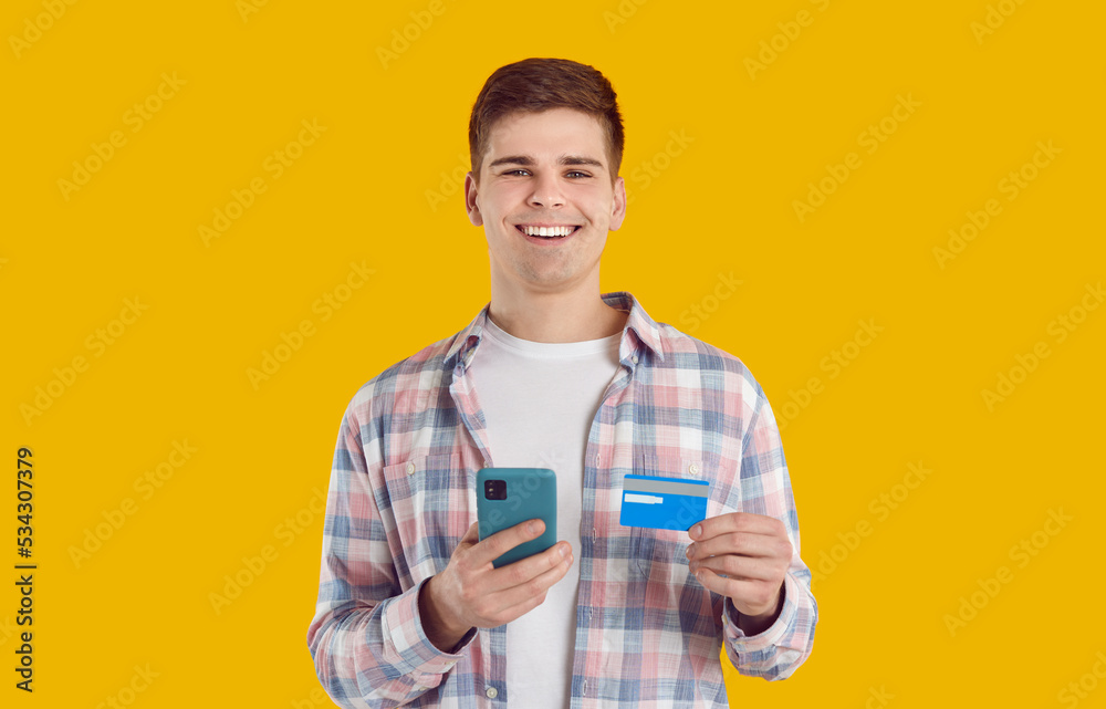 Positive young Caucasian man smiling holds phone and credit card for mobile shopping through application and payment for services on Internet sites stands on yellow studio background Positive young Caucasian man smiling holds phone and credit card for mobile shopping through application and payment for services on Internet sites stands on yellow studio background