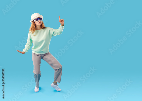 Full body length shot of funny happy pretty young woman in pastel mint sweatshirt, grey trousers, white faux fur hat and cool sunglasses dancing isolated on blue copy space background. Fashion concept