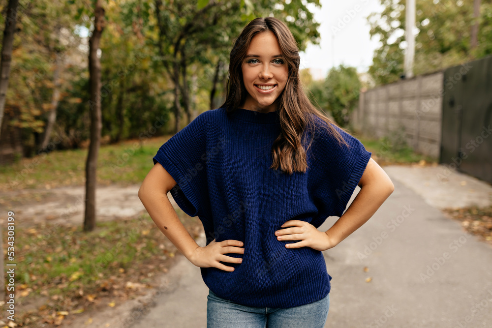 Happy exited lovely girl with wonderful smile wearing blue knitted shirt posing at camera in city on background of autumn park un sunny day.