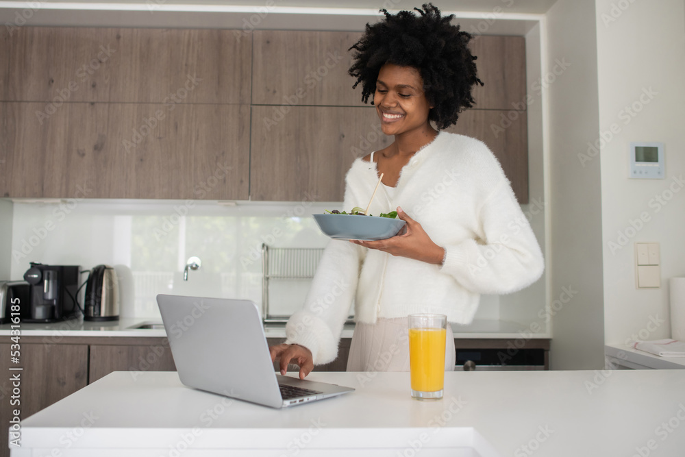 Portrait of young woman working from home typing and eating. Joyful female model typing on laptop, holding bowl of salad, standing in kitchen. Portrait, remote work concept Portrait of young woman working from home typing and eating. Joyful female model typing on laptop, holding bowl of salad, standing in kitchen. Portrait, remote work concept