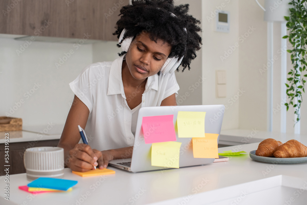 Focused woman in headphones writing on stickers. African American woman studying online during quarantine or self-isolation, sitting at table, using laptop. Online education, pandemic concept Focused woman in headphones writing on stickers. African American woman studying online during quarantine or self-isolation, sitting at table, using laptop. Online education, pandemic concept