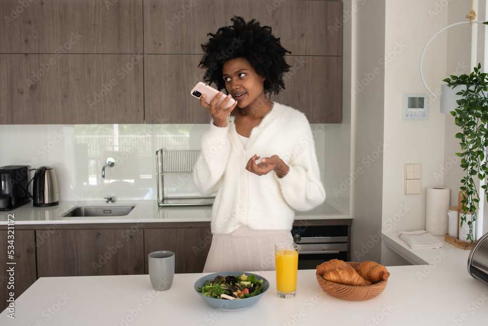 Portrait of young woman working from home sending voice message. Happy female model recording voice message to colleagues, standing in kitchen. Portrait, remote work concept Portrait of young woman working from home sending voice message. Happy female model recording voice message to colleagues, standing in kitchen. Portrait, remote work concept