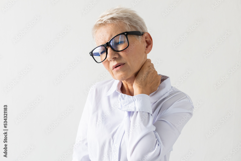 Portrait of senior woman with severe pain in neck. Female model in white shirt touching neck, rubbing, trying soothing pain. Portrait, studio shot, pain concept Portrait of senior woman with severe pain in neck. Female model in white shirt touching neck, rubbing, trying soothing pain. Portrait, studio shot, pain concept