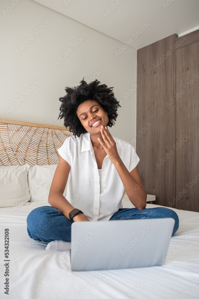 Cheerful woman talking with friends via video chat. Smiling African American woman sitting on bed, looking at laptop screen. Communication, technology, conversation concept Cheerful woman talking with friends via video chat. Smiling African American woman sitting on bed, looking at laptop screen. Communication, technology, conversation concept