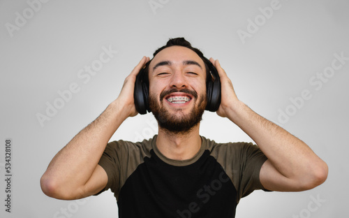 Young Latin and Hispanic man with braces and beard, happy, listening to music with his eyes closed and a smile on his face. White background.