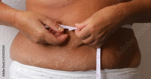 Cropped photo of naked overweight woman belly in underwear, trying to lose weight. Holding and measuring her waistline by roulette tape belly, abdomen. Fat large sagging folds on stomach. Excess skin.